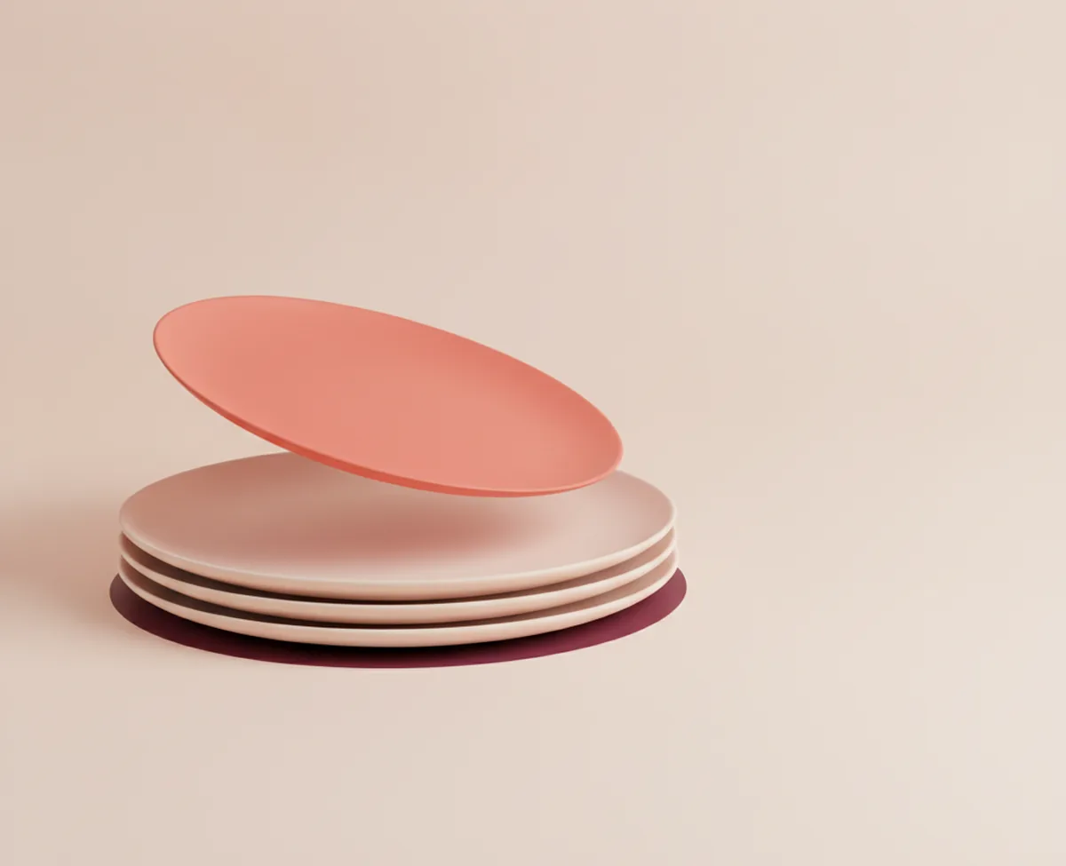 A coral disc tilted just above a neat stack of three blush discs on a warm off-white surface, a deep burgundy shadow pooling beneath the stack