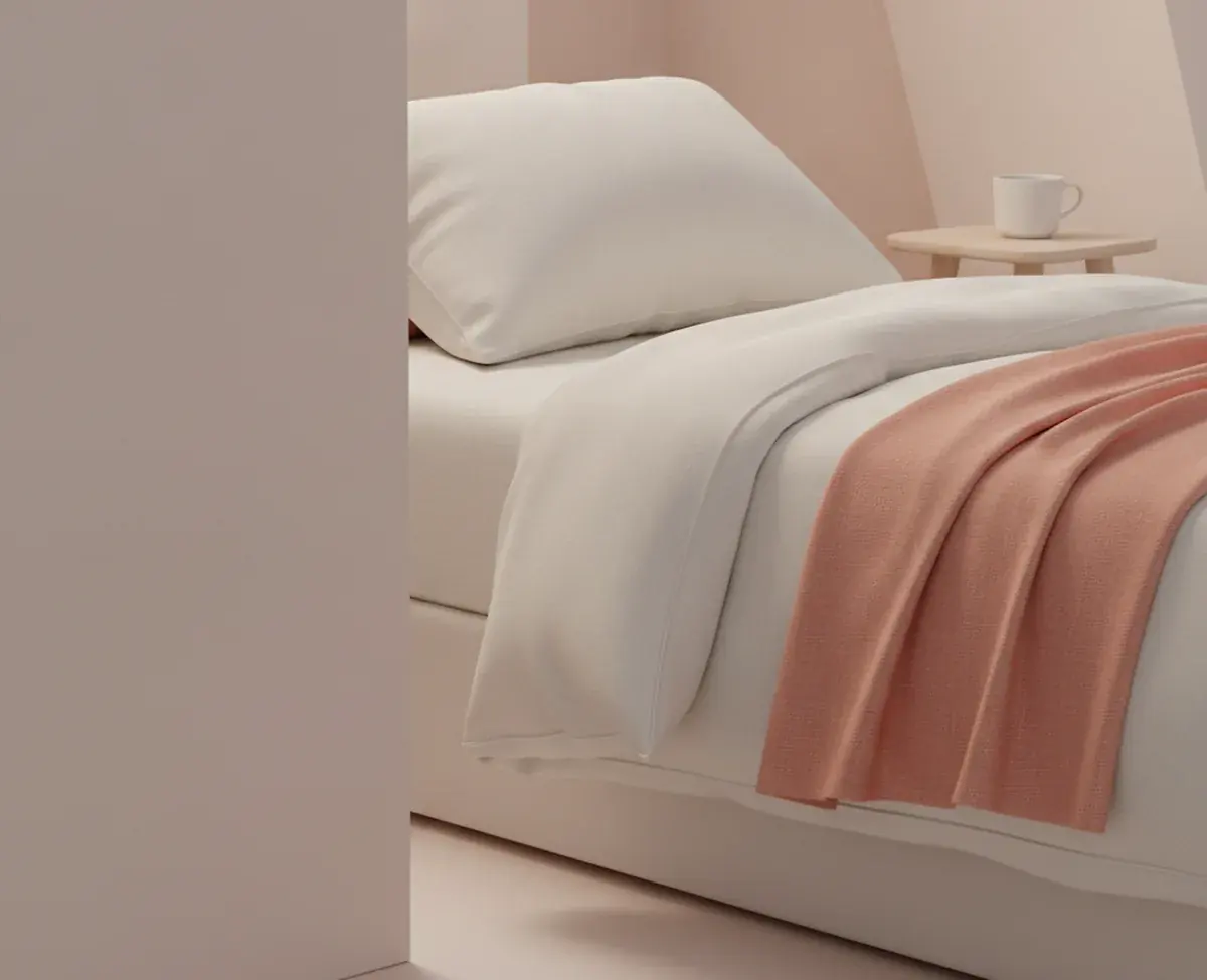The corner of a freshly made bed in morning light: crisp off-white linen and one pillow, a folded coral throw across the foot of the mattress and a plain ceramic cup on a small side table
