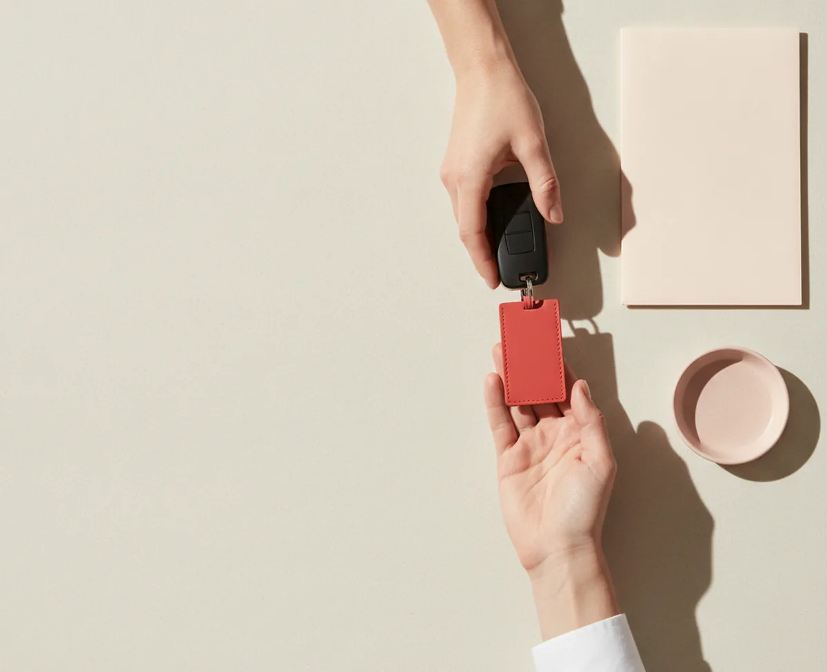 An overhead view of a pale counter where one hand passes a plain black car key on a coral leather tag to a second waiting hand, with a blank folded document wallet and an empty tray beside them