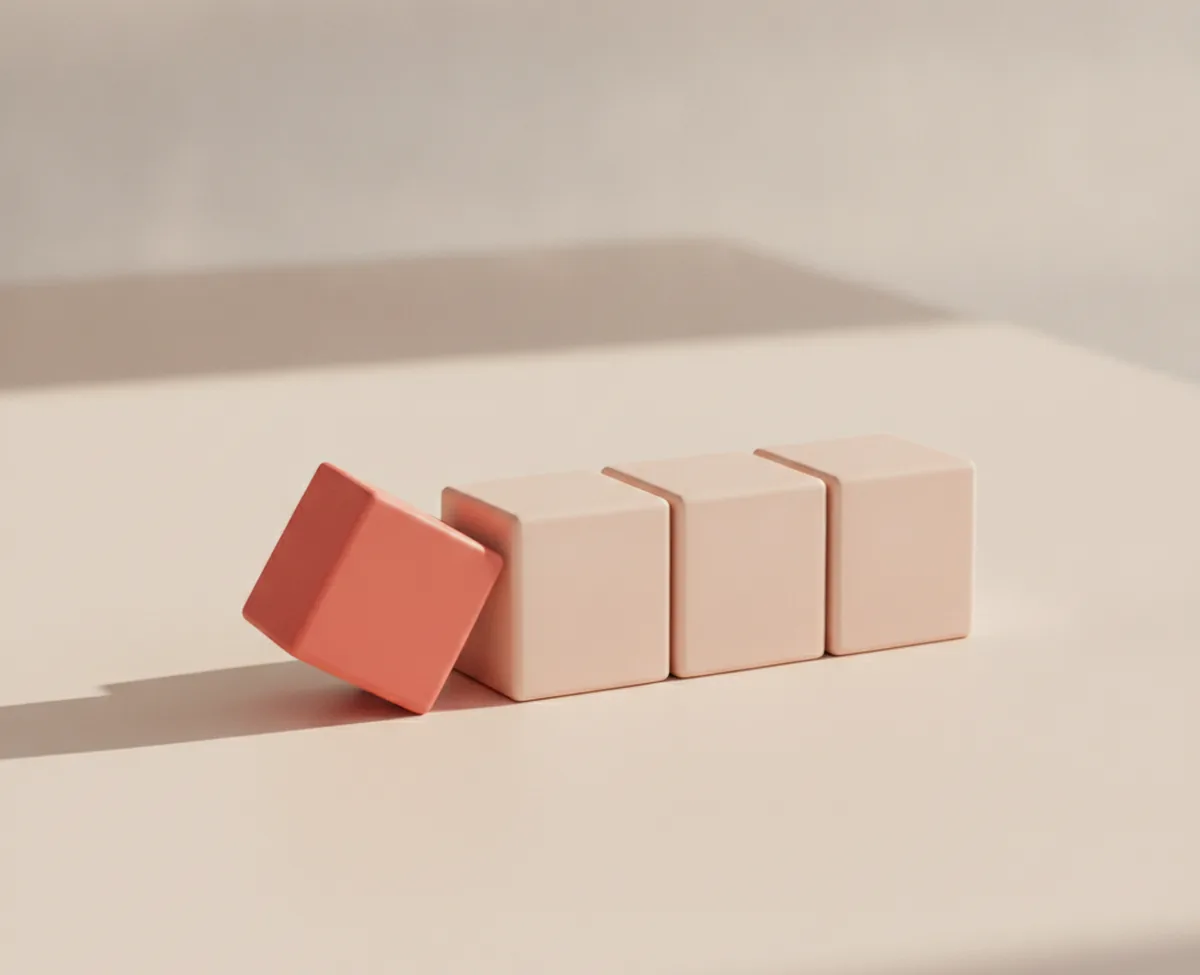 Four small matte blocks on a warm off-white surface: three blank blush cubes standing in a row and a coral one tipped over against them