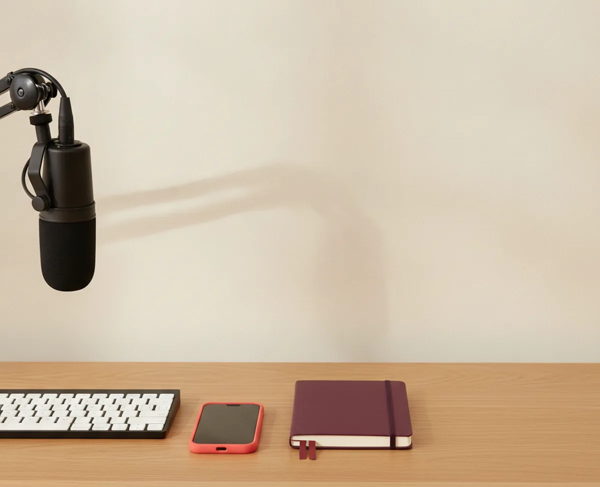 A matte black broadcast microphone on a boom arm at the edge of a warm oak desk in soft daylight, with a keyboard, a burgundy notebook and a coral-cased phone lying below it