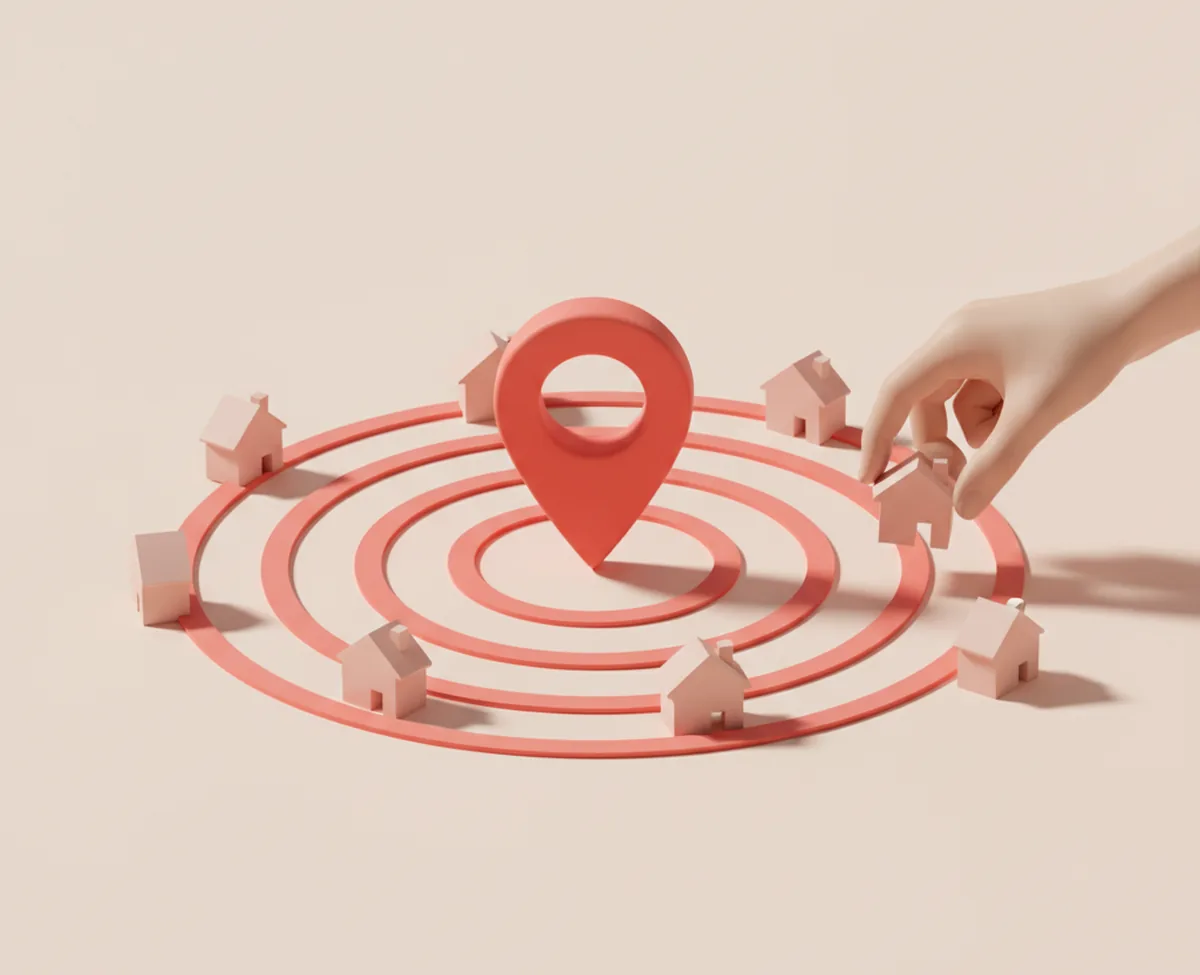 A coral map pin standing at the centre of three concentric coral rings on a warm surface, small blank houses ranged inside the rings and a hand lifting one that sits outside them