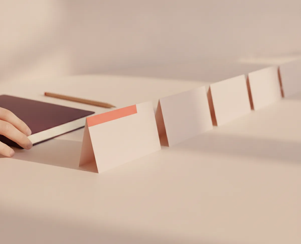 A row of blank folded cards receding across a warm off-white desk, the nearest one marked with a coral tab, a hand leaving the frame beside a closed notebook and a pencil