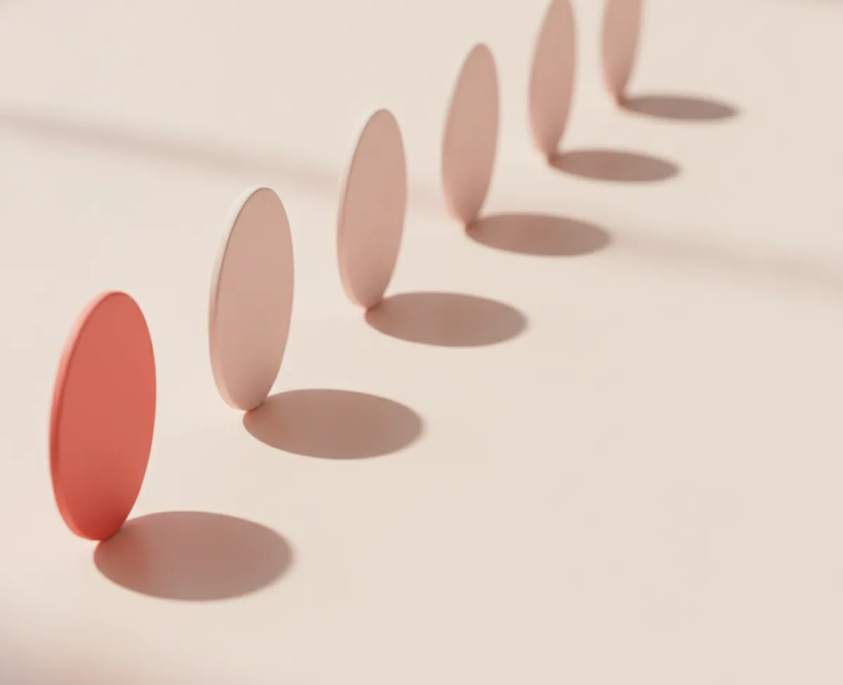 Six identical slim discs standing on edge in a receding row on a warm off-white floor, the nearest one coral and the rest pale blush, each casting the same soft shadow at the same interval