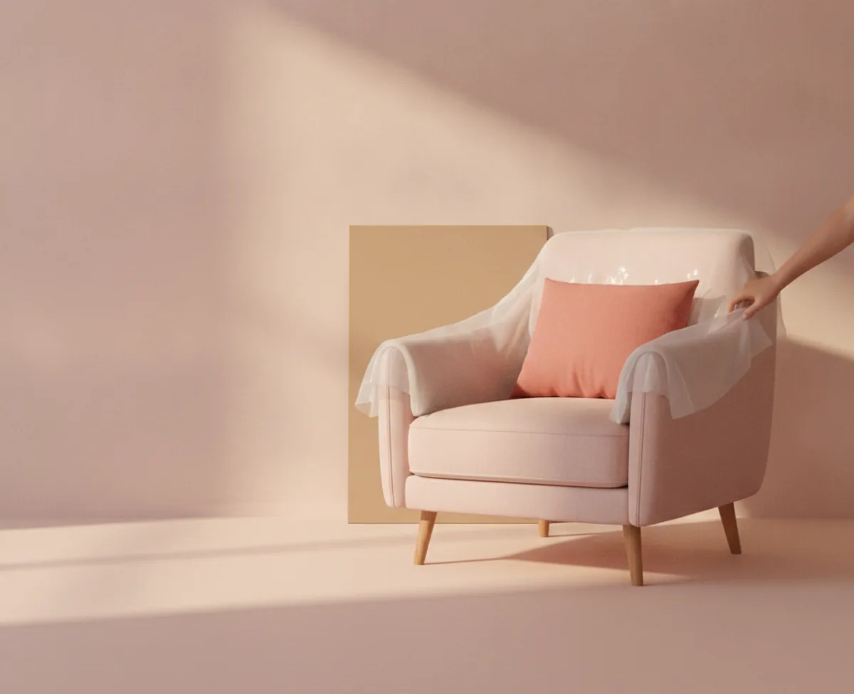 A blush upholstered armchair standing in an otherwise empty room, still half covered in clear protective wrapping, with a hand drawing the wrapping back and a coral cushion on the seat