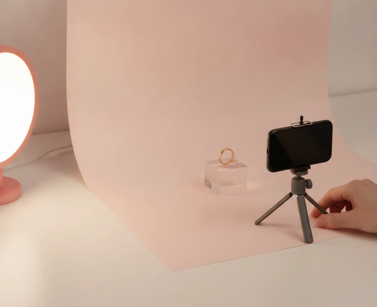 A small tabletop photo set on a desk: a blush paper sweep with a plain gold band ring on a clear riser, a phone on a tripod with a blank screen aimed at it and a coral lamp at the edge of frame