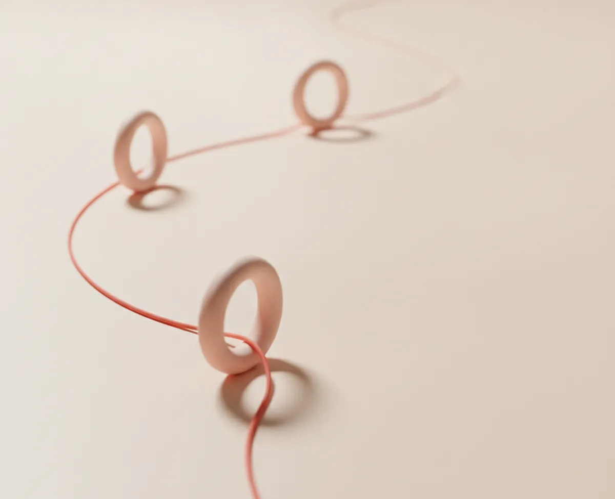 A coral thread curving across a warm off-white surface and passing through three upright blush rings standing along its path, the nearest ring in sharp focus
