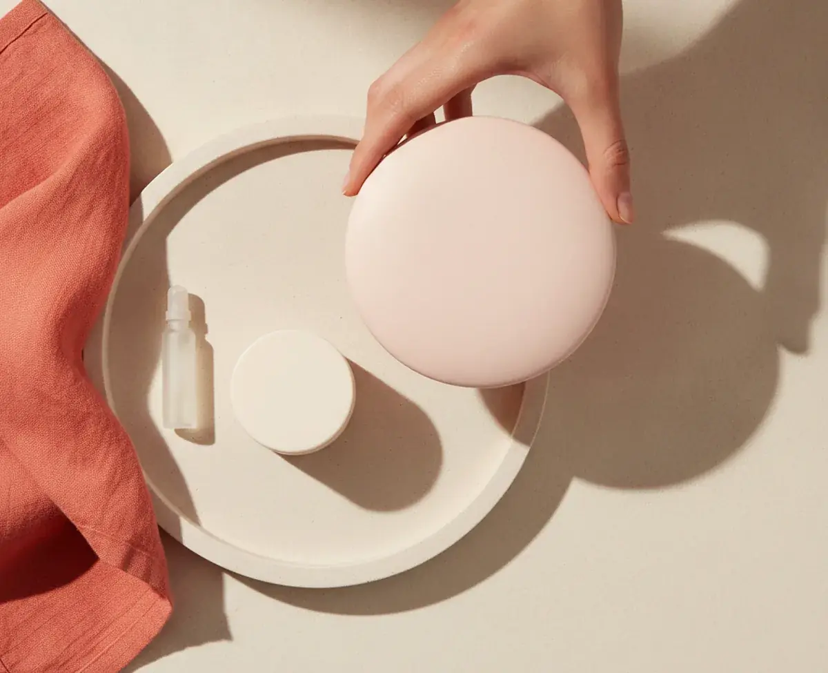An overhead of a pale stone tray holding a tiny unlabelled vial and a small cream jar, with a hand lifting a much larger plain blush beauty device clear of the tray beside a coral linen cloth